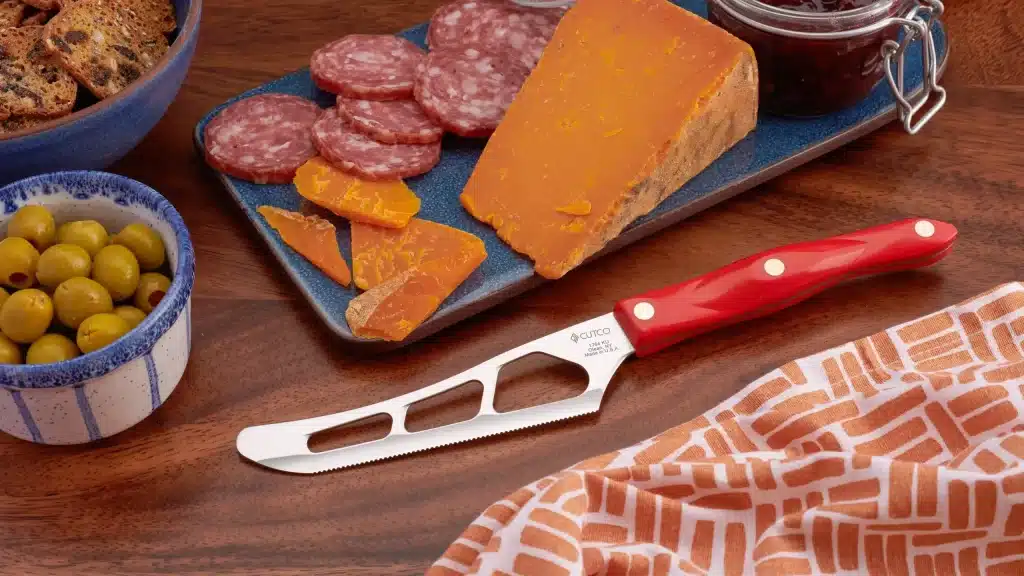 Cheese knife with red handle displayed beside cheddar cheese, salami, olives, and crackers